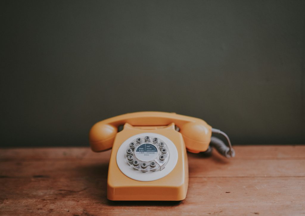 alt="An orange rotary-style phone sits on a wooden table. It has push buttons instead of the typical rotary mechanism."