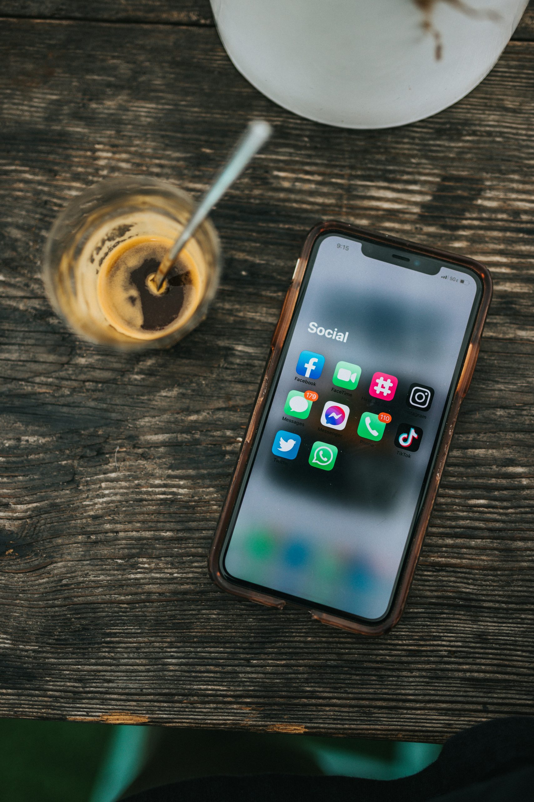 alt="An iPhone sits next to a nearly-finished coffee. The home page is open to a folder of social media apps, including Facebook, Instagram, TikTok, Twitter, and other messaging apps."